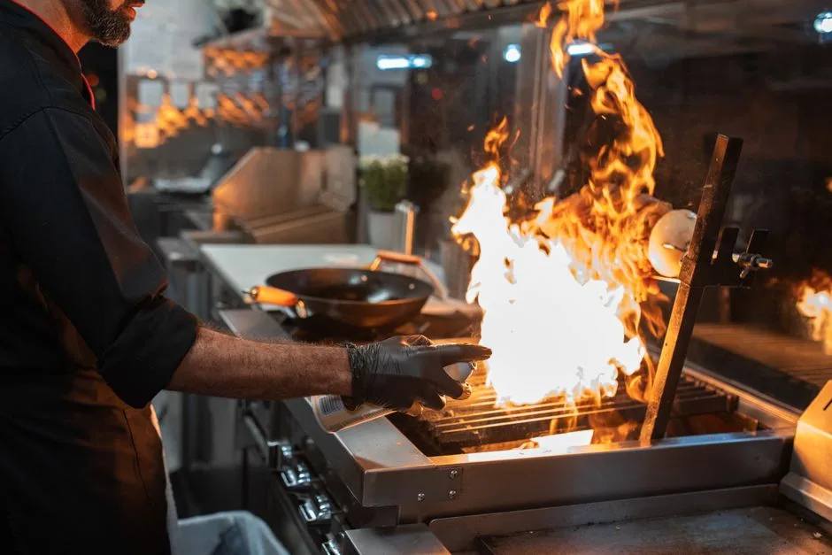 Chef grilling meat with open flames in a Hammonton restaurant kitchen — grease and smoke captured by overhead exhaust hood